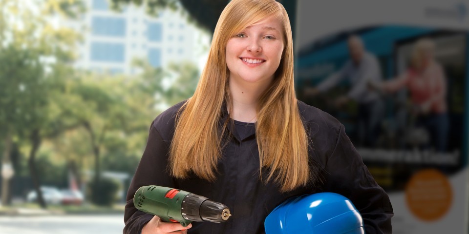 Eine junge, freundlich lächelnde Frau mit langen, blonden Haaren und schwarzem Oberteil hält eine Bohrmaschine und einen Schutzhelm in den Händen und blickt in die Kamera.