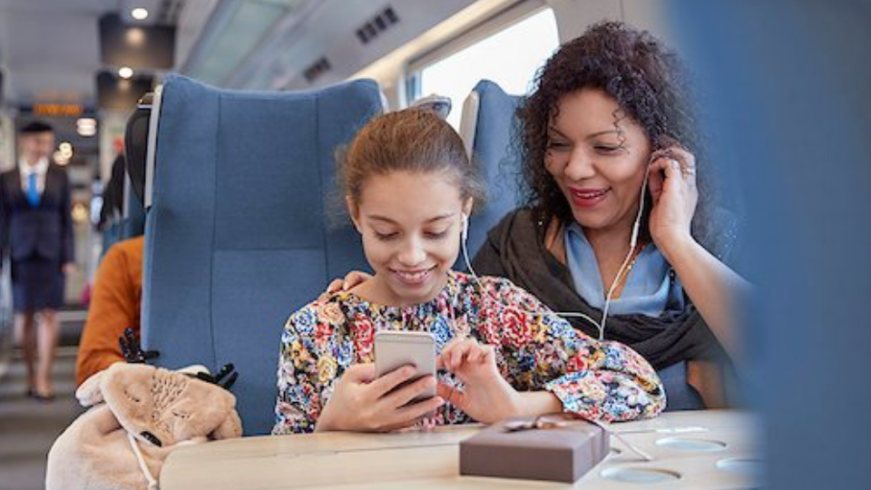 Mutter und Tochter sitzen im Zug und blicken lächelnd gemeinsam auf ein Smartphone, beide tragen je einen Kopfhörer im Ohr.